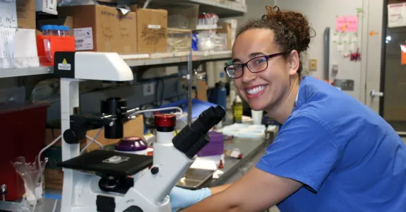 Indoor photo of a Black female graduate student working at a microsocope in a laboratory.