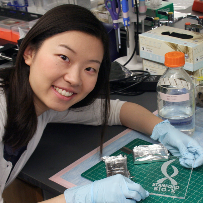 Photo of graduate student Kiara Cui in the lab, holding a glass plate with the Bio-X logo laser-cut into the surface.