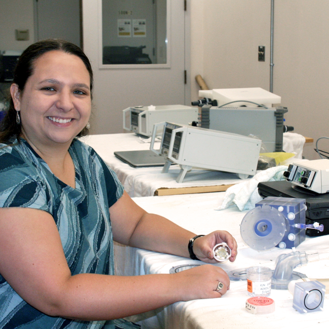 Photo of graduate student Nicole Schiavone in a dry product design lab space, holding up small plastic prototypes of a heart valve repair device.