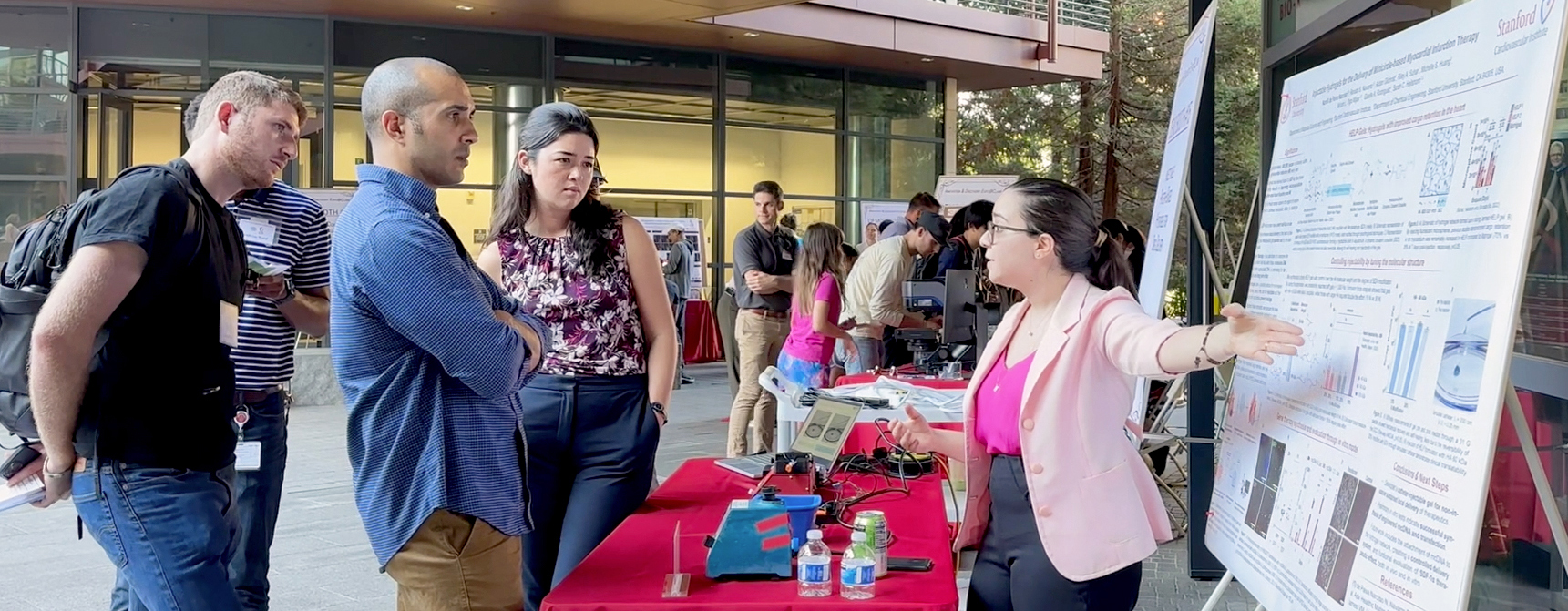 Outdoor photo of a young female researcher displaying a poster to other researchers next to a table with devices on it.