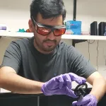 Indoor photo of a graduate student wearing orange lens safety goggles holding up a device.