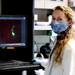 Indoor photo of a graduate student standign next to a computer displaying images of cells.