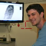 Indoor photo of a graduate student standing next to a monitor screen displaying a cell.