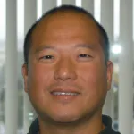 Indoor headshot photo of a smiling Asian male faculty member, Dr. Stuart Kim, Professor Emeritus of Developmental Biology at Stanford University.