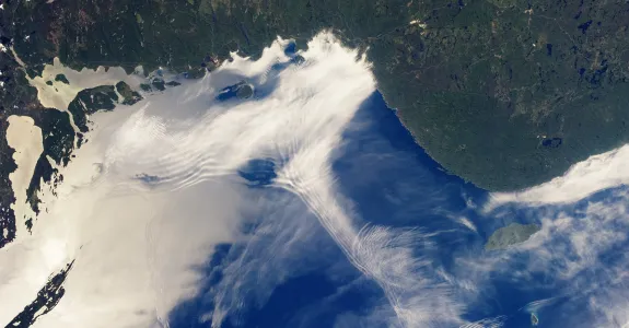 Satellite image of a large body of water and the nearby land, with numerous clouds rippling in different ways.