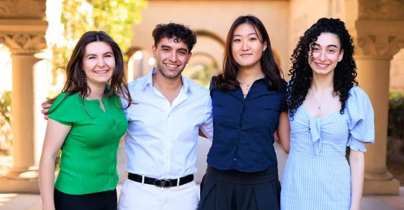 Outdoor photo of 4 undergraduate students standing together, smiling at the camera with their arms around each other.