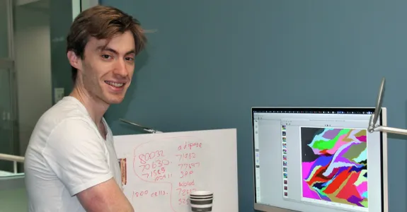 Photo of white male graduate student standing next to a computer displaying colorful data on a large screen.