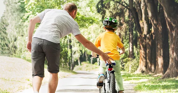Photo of a father helping his son to learn to ride a bike in a wooded area.