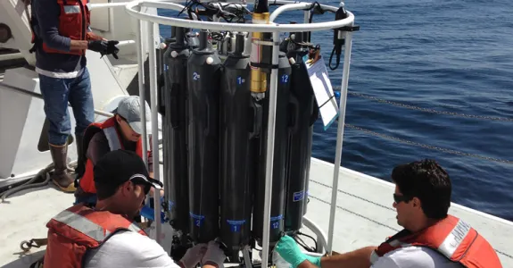 Photo of two men in orange life jackets attending to a sample collector, which is large and cylindrical in shape and comprised of multiple black collection tanks.