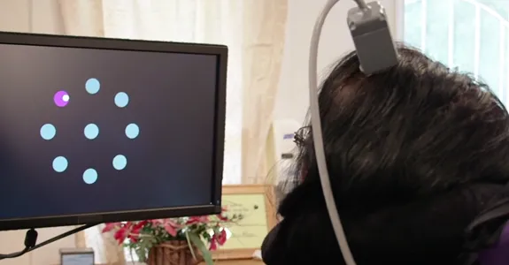 Screenshot from video showing paralyzed woman with device attached to head, using neural signals to manipulate cursor on a screen to select dots lighting up.