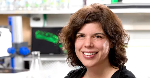 Photo of smiling white female faculty member with shoulder-length wavy brown hair, with a wet lab in the background behind her.