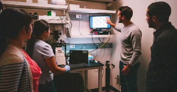 Indoor photo of four diverse lab members gathered around a computer screen, with one male graduate student gesturing to the display.