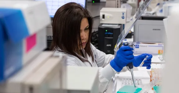 Photo of a female researcher in a lab coat and gloves pipetting into tubes in a laboratory area.