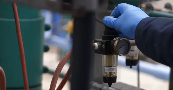 Screenshot/image of a man's hand in a blue glove adjusting a valve on a pipe.