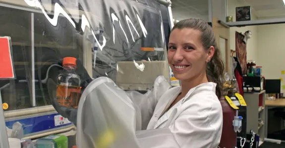 Indoor photo of a white female graduate student with her arms put into a safety hood with plastic, holding up a bottle inside the hood, and smiling at the camera.
