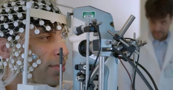 Photo of a man with a net of electrodes on his head with his face resting on a chin rest in front of a machine, with a professor in a lab coat watching from the background.