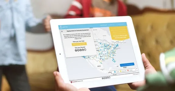 Photo of person holding iPad, with screen displaying GapMap tool, while two children play in the background.