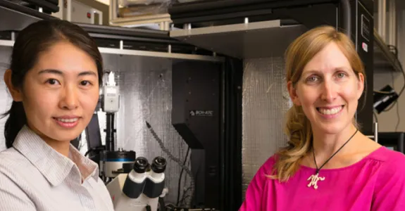 Photo of a young Asian woman and her female Principal Investigator, standing in a specialized lab space with a microscope.