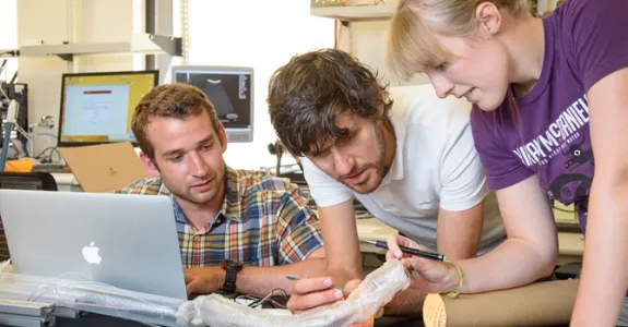 Male and female graduate student work with male visiting professor, consulting a computer screen and examining the translucent "vinebot" robot device.