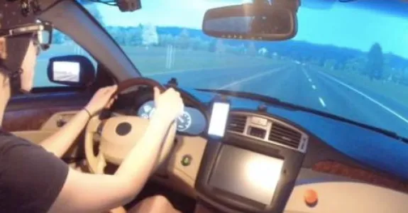 Photo of a young male study participant sitting in a car with a simulated street image past the windshield; participant is wearing a black cap with wires trailing from it and has both hands on the steering wheel.
