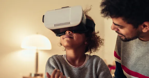 Photo of a young African American girl wearing a VR headset, sitting down with her father.