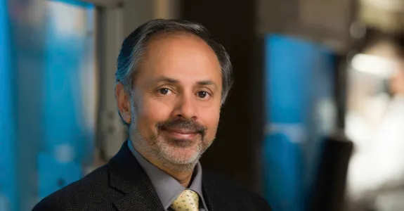 Photo of a smiling man of Indian descent with a beard, wearing a suit and sitting in a laboratory area.