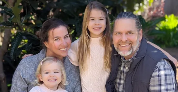 Photo of woman and man with their two young daughters, posed in front of trees.