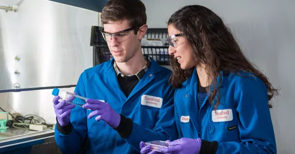 Photo of students Colin McKinlay and Jessica Vargas in the laboratory, examining a test tube and a petri dish near a fume hood.