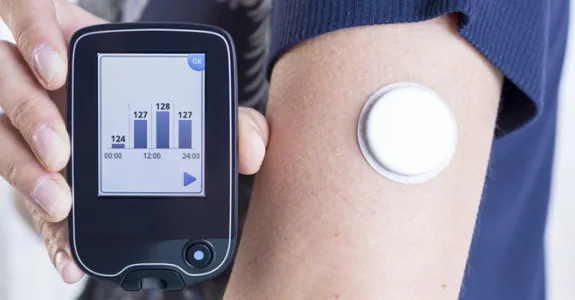 Photo of a woman's arm which bears a glucose monitor, holding up a readout.