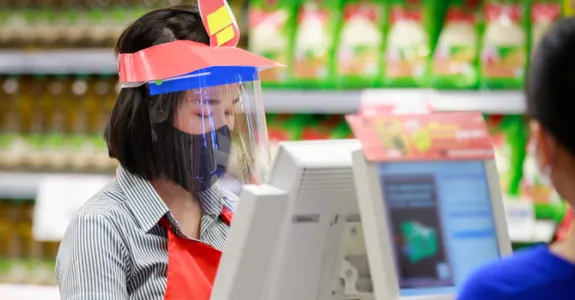 Photo of a female grocery store cashier wearing a mask and a face shield, checking out a shopper wearing a mask.