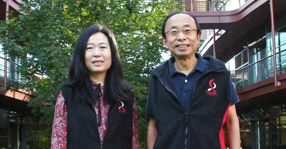 Outdoor photo of an Asian female faculty member and Asian male faculty member smiling at the camera outside of the Clark Center, wearing matching fleece vests with the Bio-X logo.