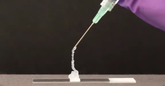 Photograph of a needle extruding a pale translucent gel onto a glass slide, with the photo centered on the needle and showing just a sliver of a purple gloved hand holding it above the slide.