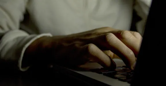Photo of a man using a computer in the dark, showing only his hand on the keyboard and not his face.