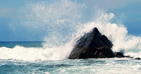 Photo of ocean waves crashing against a large rock and spraying.