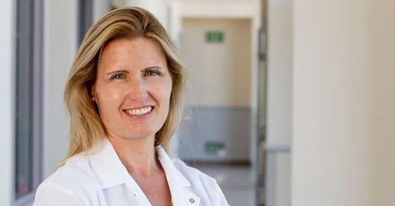 Photo of blonde female PI standing in a hall, smiling, while wearing a white coat.