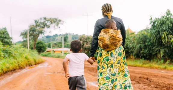 Photo of Kenyan woman walking down a dirt road, with one small child carried in a sling on her back, holding the hand of a slightly older child.