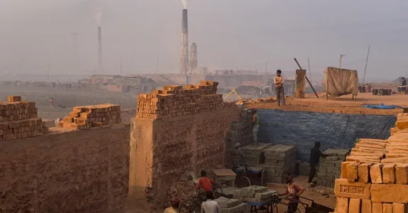 Photo showing workers moving bricks, surrounded by walls made of other bricks, with kilns smoking in the background.