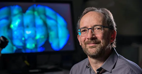 Photo of Dr. Mark Krasnow in lab, next to microscope and in front of a screen displaying a large brain scan image in bright blue.