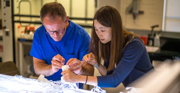 Photo of a male and female scientist both leaned over a plastic device on an illuminated table, adding wires to it.