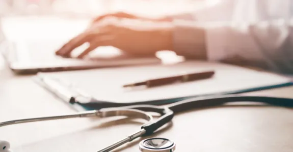 Photo of a doctor's hands using a laptop in the background, with a stethoscope on the table in the foreground.