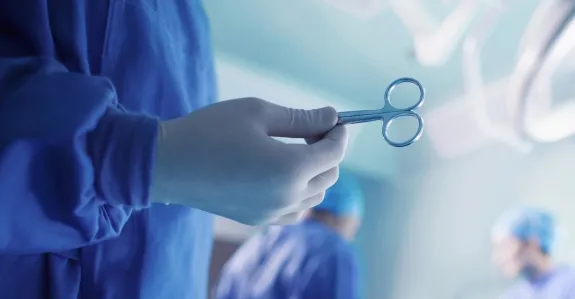 Photo of a surgeon's gloved hand holding up a pair of surgical scissors in the foreground, with operating room activity in the background.