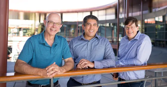 Outdoor photo of Scott Delp, Gabriel Sanchez, and Mark Schnitzer  standing at a railing on an upper level of the Clark Center at Stanford.