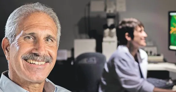 Photo of a middle-aged male PI with a mustache, with a student working on neuroscience displays in the background.