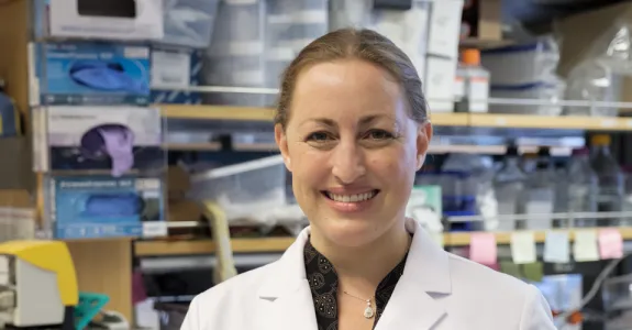Photo of smiling female faculty member, Dr. Michelle Monje, standing in a wet laboratory space wearing a lab coat. Her blond hair is tied back.