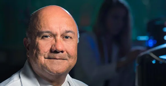 Photo of Dr. Jose Montoya in the laboratory, wearing a white coat, with a researcher working in the background.