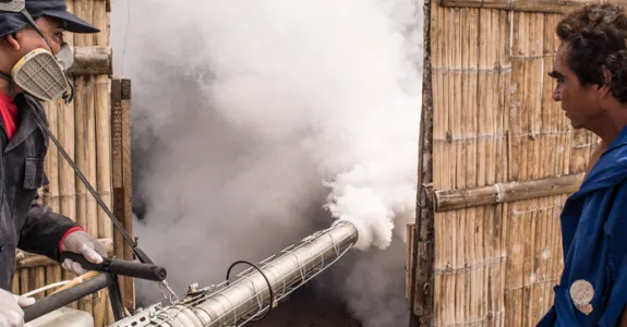 Photo of man in safety gear spraying smoke into a enclosed fence area while another man looks on.