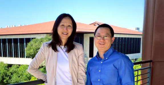 Outdoor photo on a balcony of a smiling Asian female faculty member wearing a pink blazer and a white shirt, with long black hair, and a slightly shorter, smiling male Asian faculty member wearing glasses and a royal blue button up shirt. In the distance behind them are bright green trees and a building with many windows and a red brick roof.