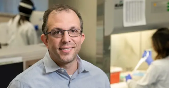 Photo of male faculty member with glasses standing in the lab, with a researcher using a fume hood in the background.