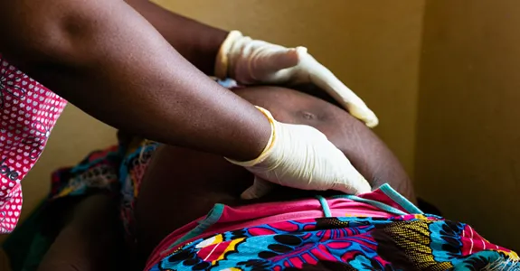 Photo of an African doctor wearing pink scrubs and pale yellow gloves examinign a pregnant African woman.