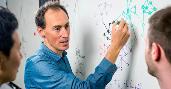 Photo of Dr. Jonathan Pritchard (center) and his colleagues, Yang Li (left) and Evan Boyle, with Pritchard having drawn several interconnecting gene trees and formulas on a whiteboard in different colors.
